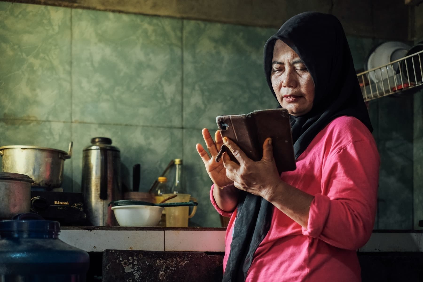 Foto Ibu Warni memegang ponselnya dan berada di dapur.