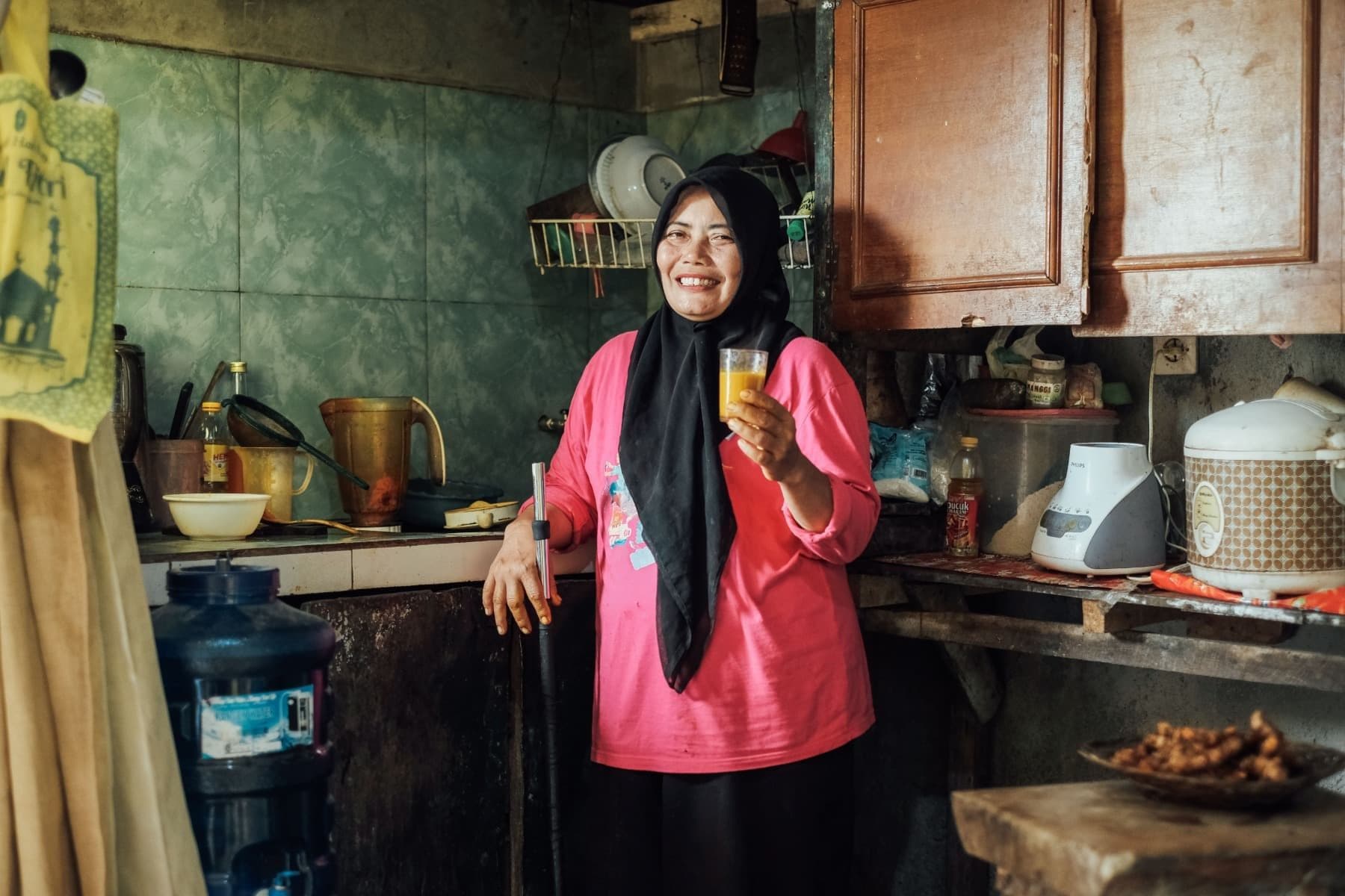 Foto Ibu Warni sedang tersenyum sambil memegang segelas jamu buatannya di dapur.