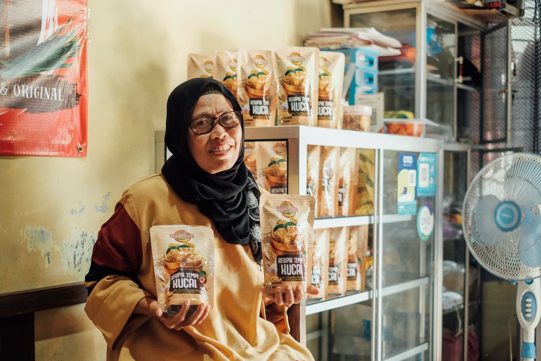 Foto Ibu Rustini tersenyum sembari membawa produk Keripik Masshita di kedua tangannya.