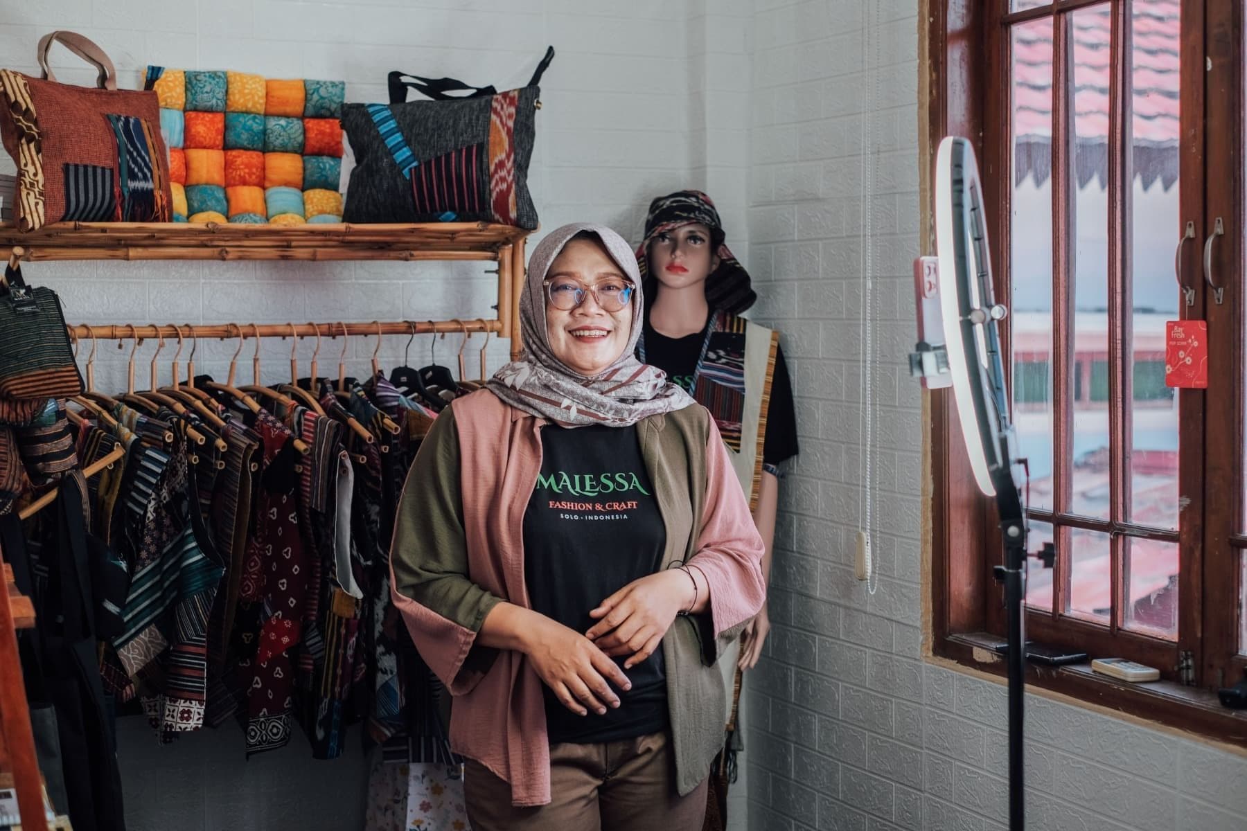Foto Ibu Madu berdiri di depan rak yang dipenuhi oleh produknya sembari tersenyum.