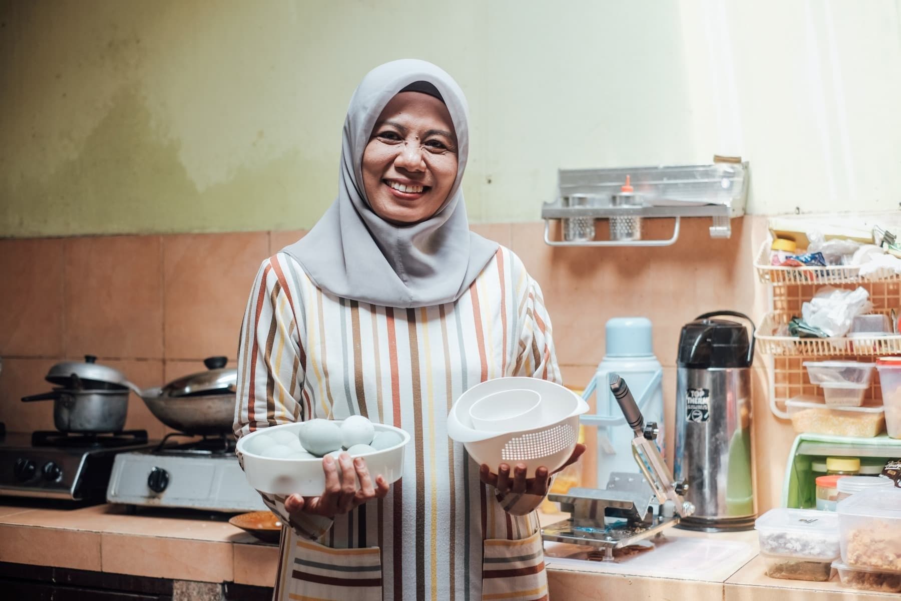 Foto Ibu Ririn tersenyum sambil memegang telur dan berada di dapur.
