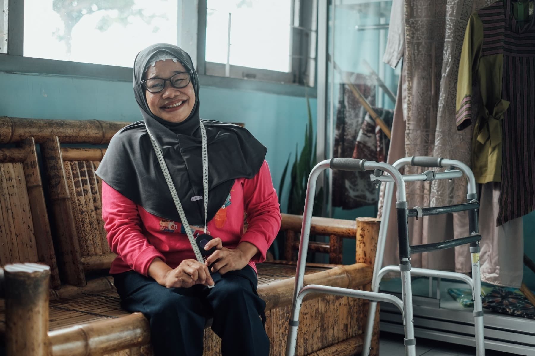 Foto Ibu Endang tertawa sambil duduk di kursi bambu, dengan meteran jahit di lehernya dan alat bantu jalan di sampingnya.